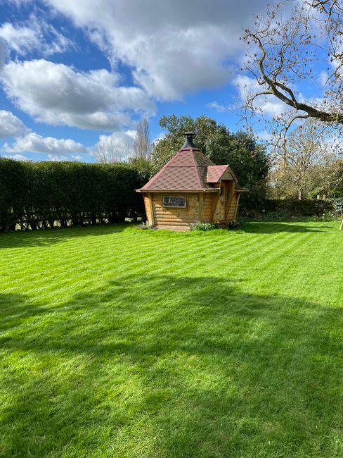 A well-maintained garden with a neatly mowed lawn featuring lush, bright green grass with visible mowing stripes. In the background, there is a small wooden garden shed with a red, pitched roof and a 